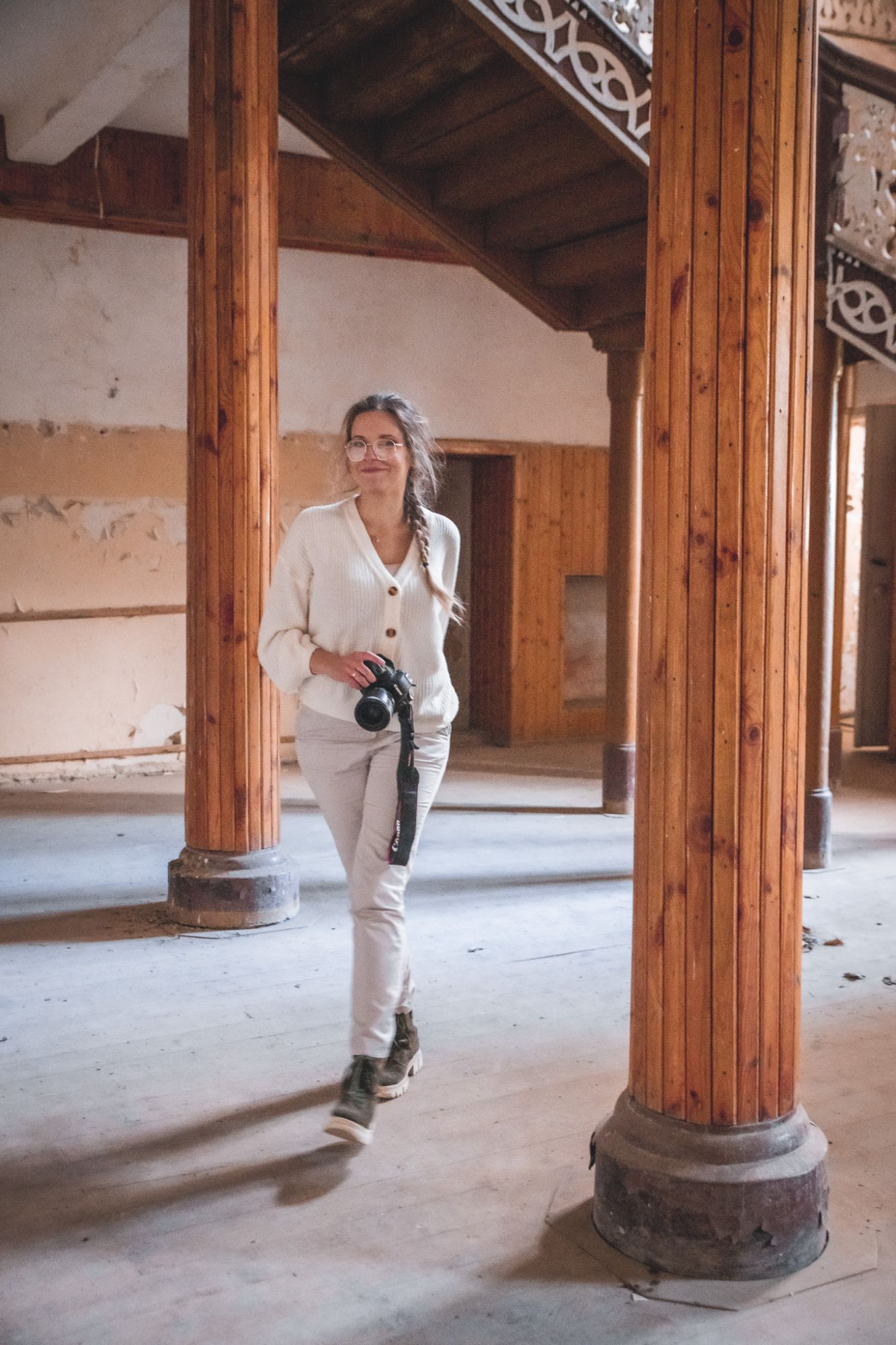 Kobieta w białym swetrze, trzymająca aparat fotograficzny, spaceruje po starym wnętrzu opuszczonego pałacu